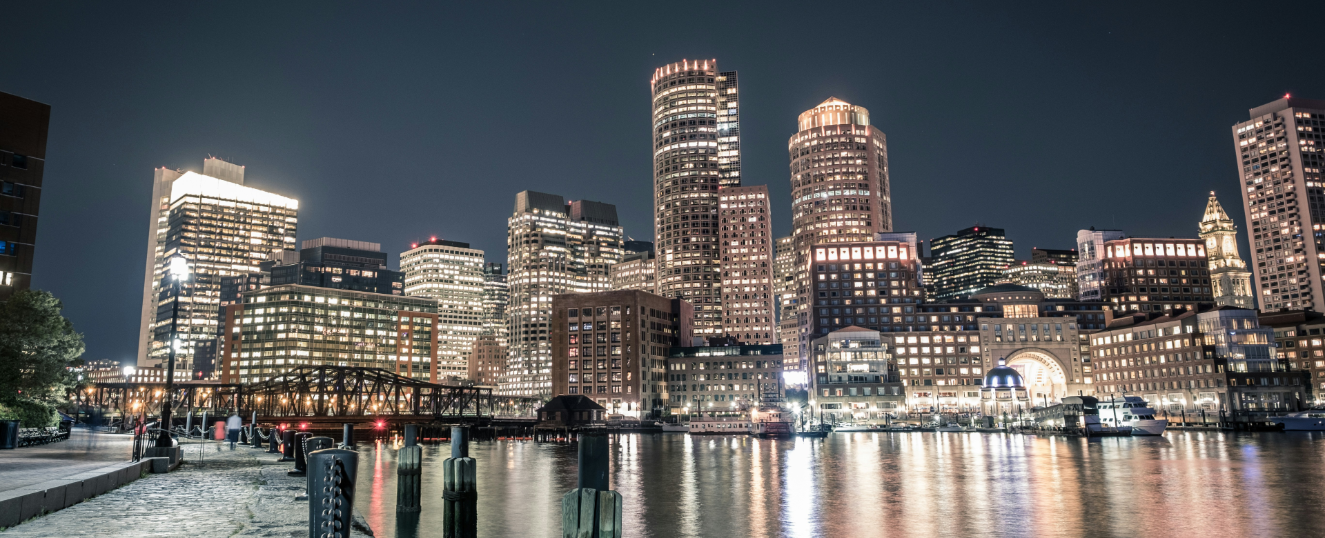It's a night view of Boston, Massachusetts. The distinctive skyline, including the Prudential Tower and the Custom House Tower, along with the waterfront and the Charles River, are clear indicators of the city.