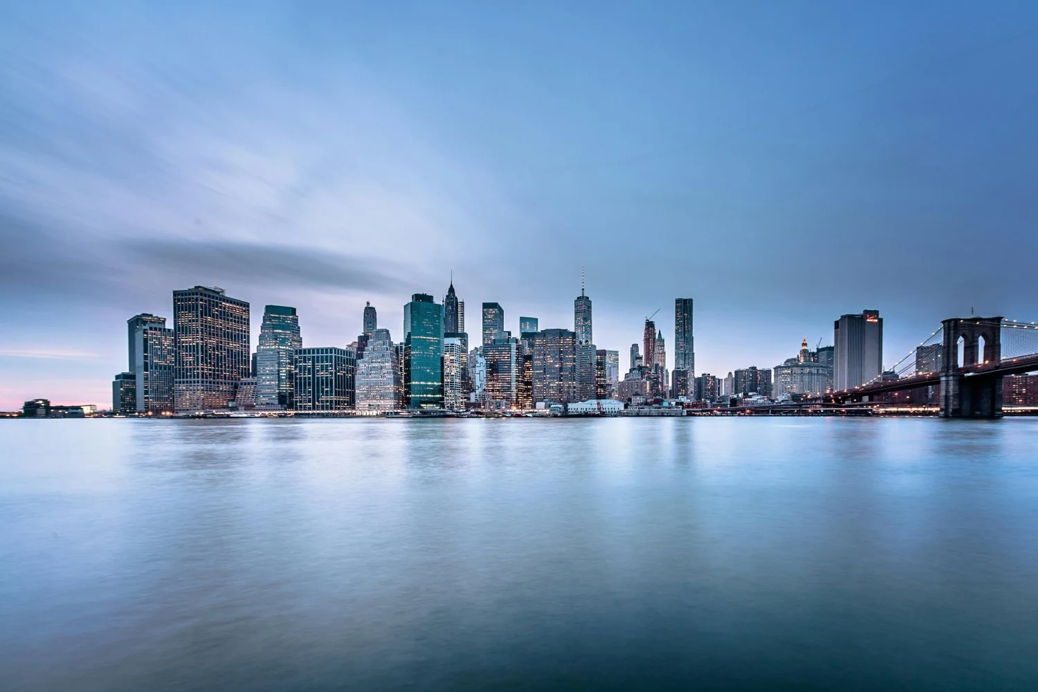 New York City skyline at dusk. The Brooklyn Bridge is visible in the foreground, spanning the East River.