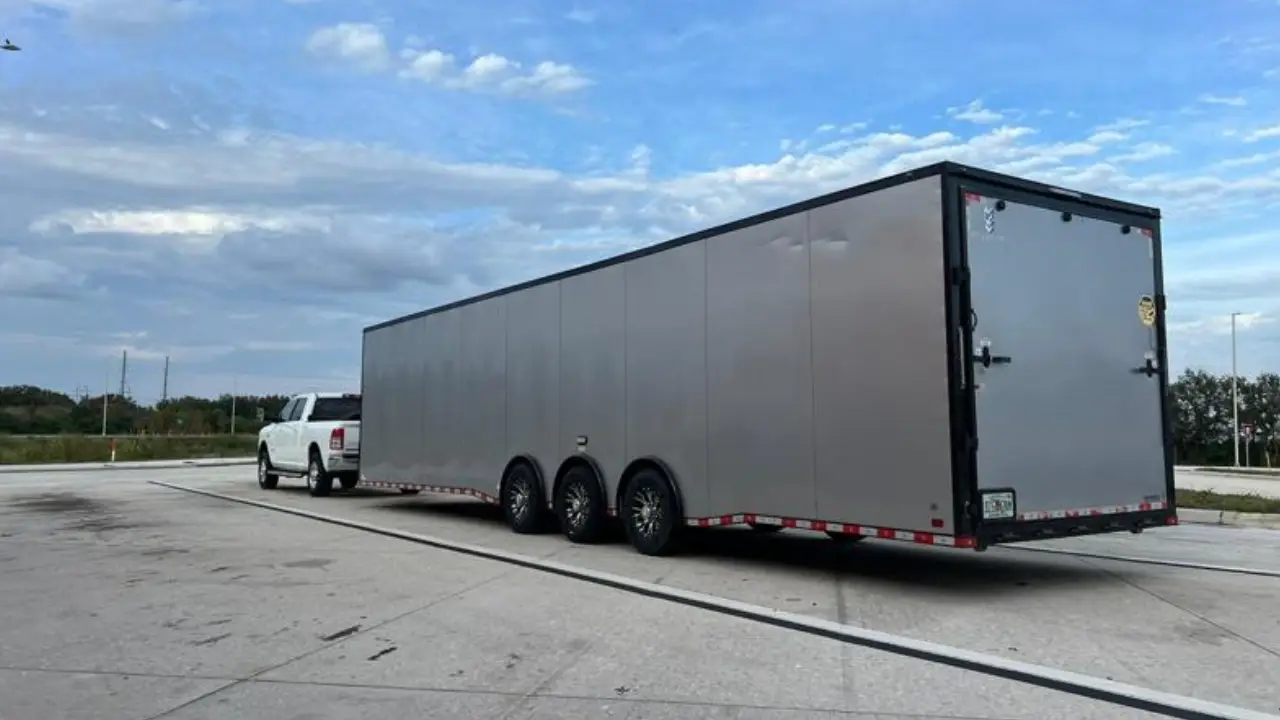 A white pickup truck towing a very long, multi-axle, silver enclosed trailer under a cloudy sky